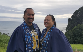 Two people in graduation attire smiling. In the background is an ocean and trees.